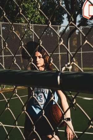 Basketball player seen through chain link fence holding ball on outdoor court.