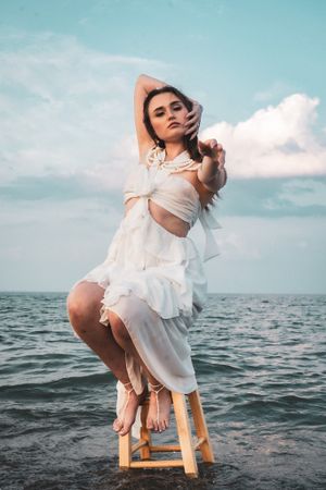 Artistic portrait of figure in white dress posing on stool in ocean water.