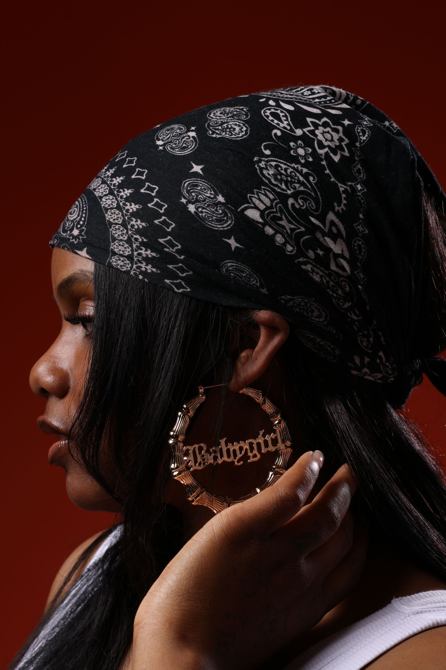 Woman in black bandana headscarf wearing large gold hoop earrings reading Babygirl, profile view on red background.