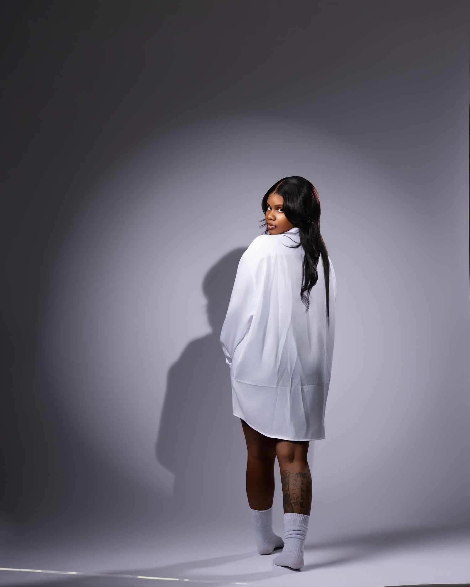 Woman in oversized white hoodie dress and white socks poses with back turned in a studio setting.