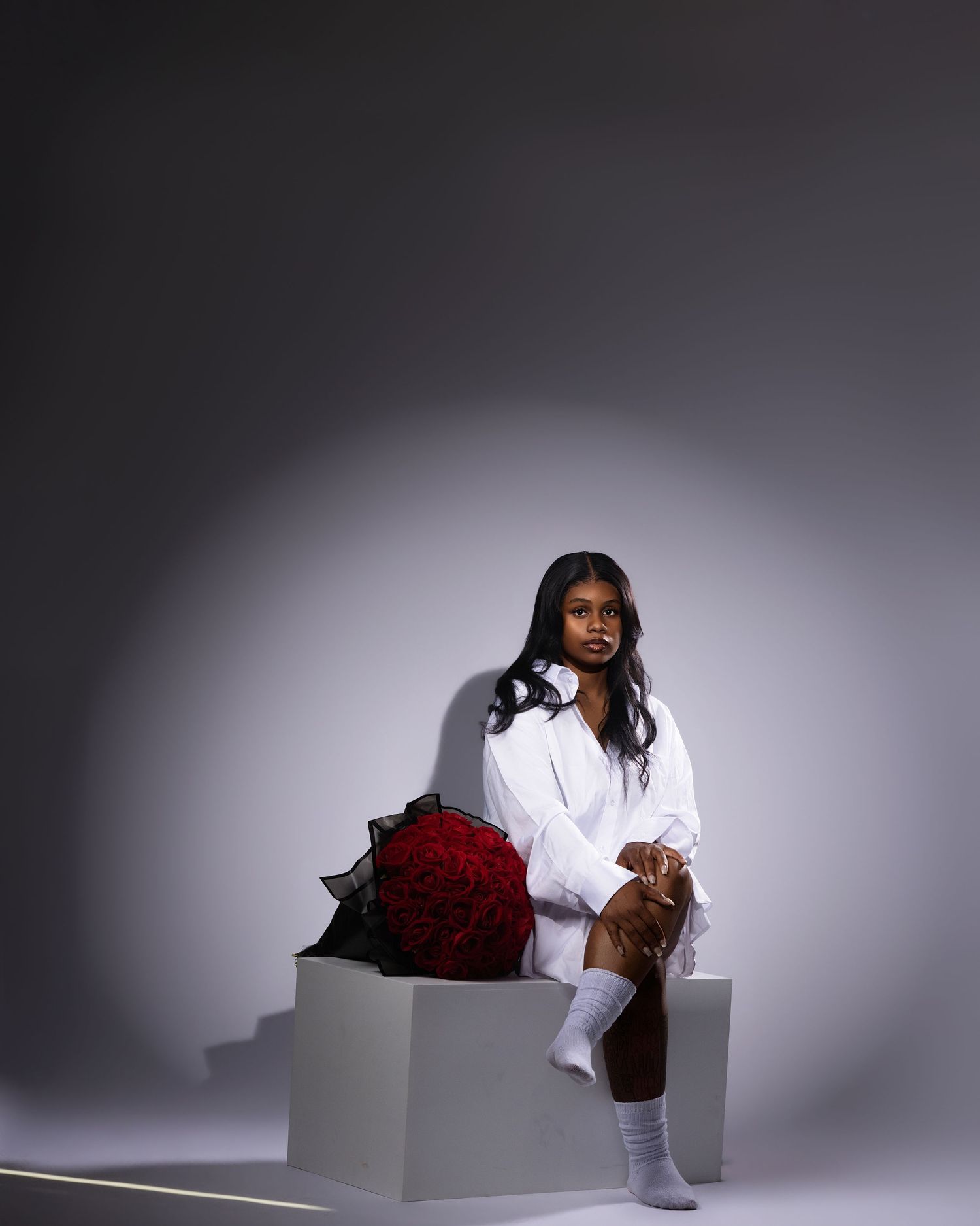 Young woman in white oversized shirt and socks sits on white cube beside large red rose bouquet against gray background.