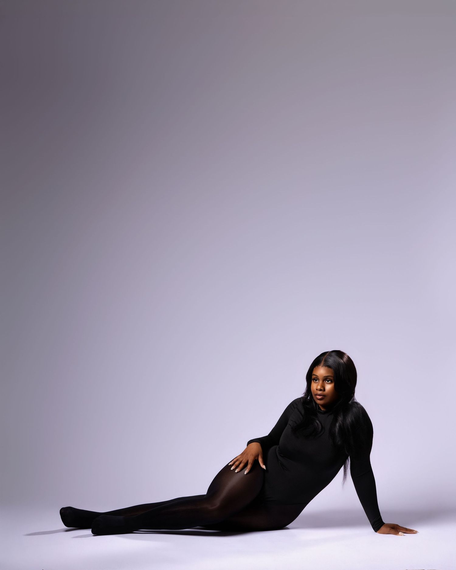 A Black woman in a black bodysuit and tights poses gracefully on a white studio floor.