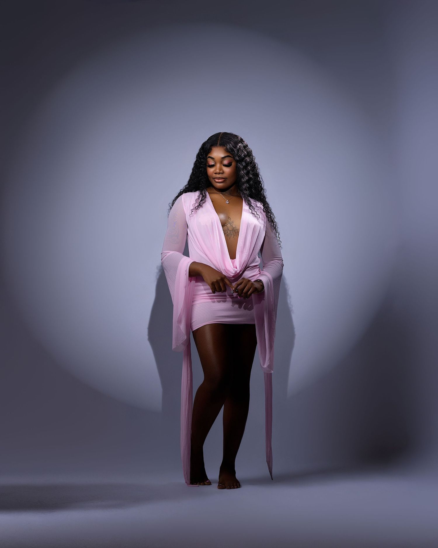 Woman in pink mini dress with sheer sleeves posing confidently against a gray studio background.