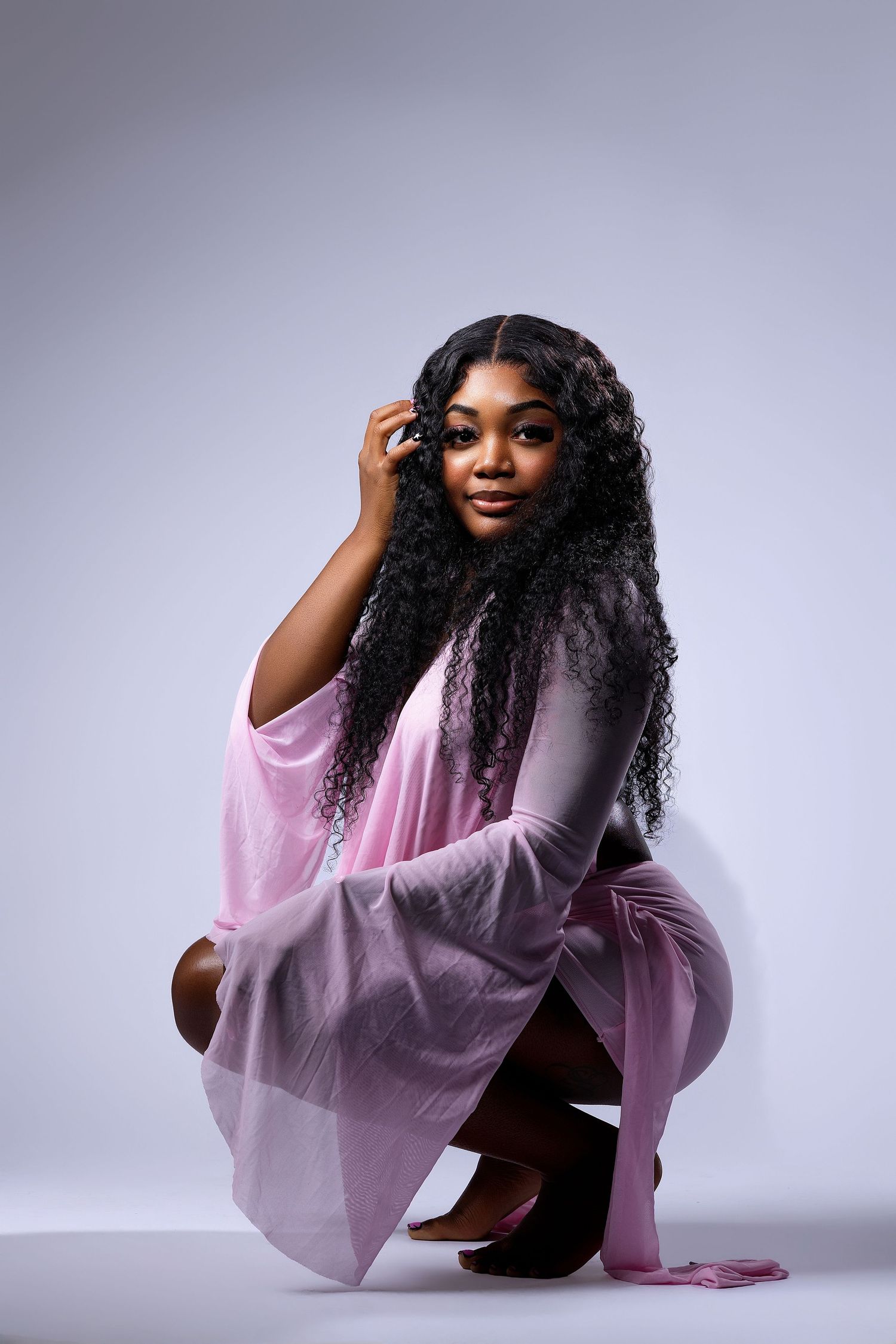 Young Black woman in pink velvet outfit with long curly hair posing gracefully in a studio setting.