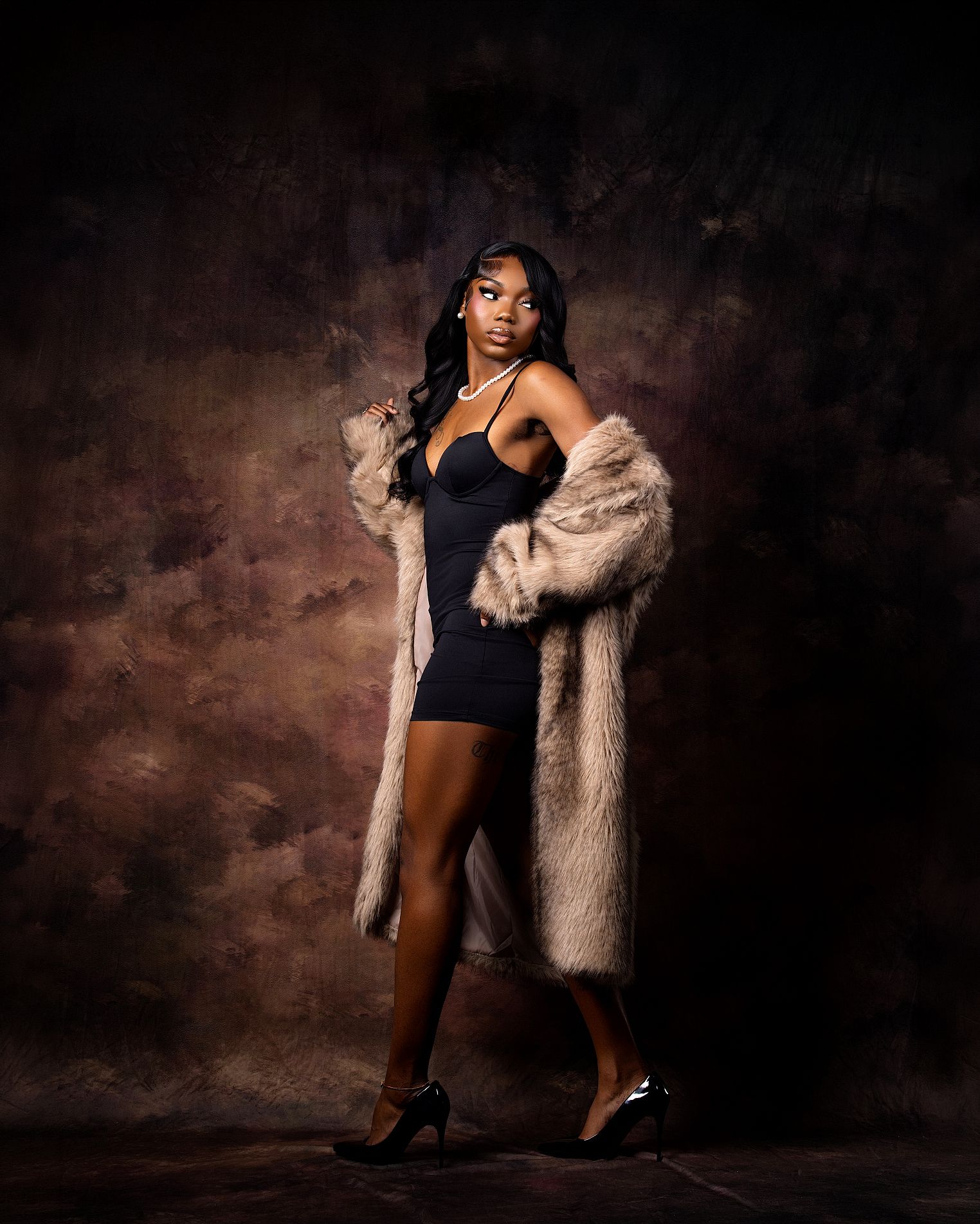 Woman in black mini dress and fur coat poses elegantly against dark studio backdrop wearing pearl necklace and heels.