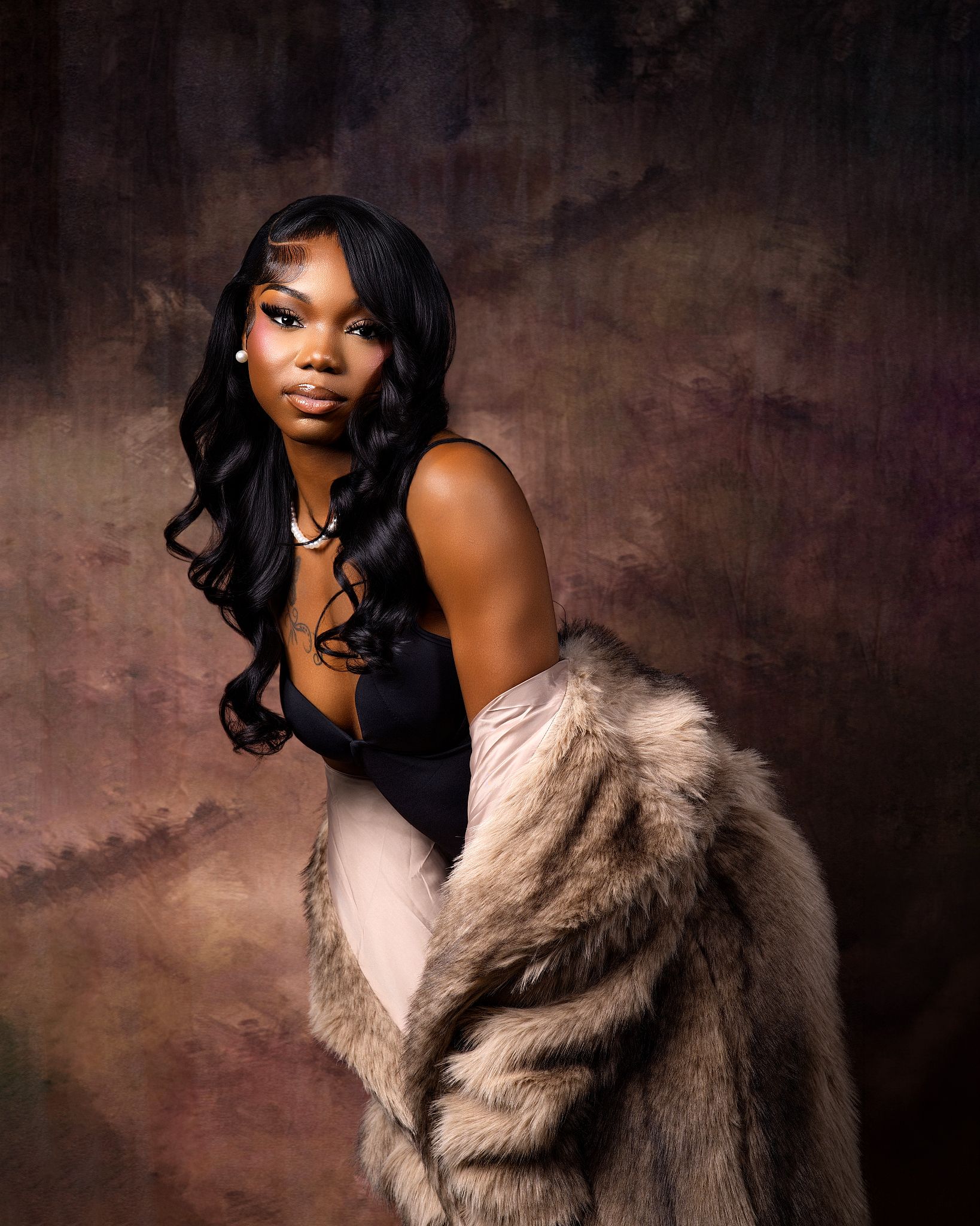 A young Black woman poses elegantly in a black bodysuit and fur coat against a moody brown studio backdrop.