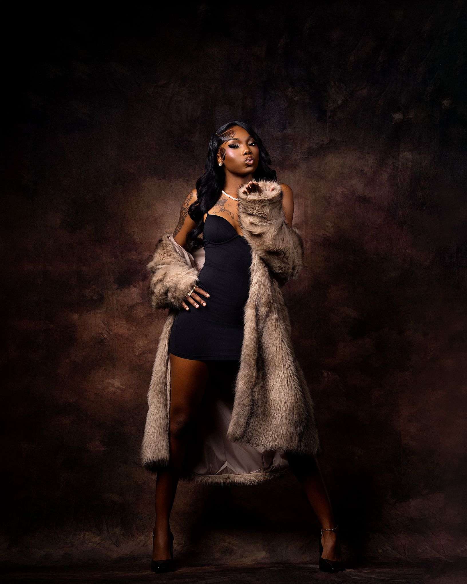 A woman in a black mini dress and fur coat poses confidently against a dark studio backdrop.