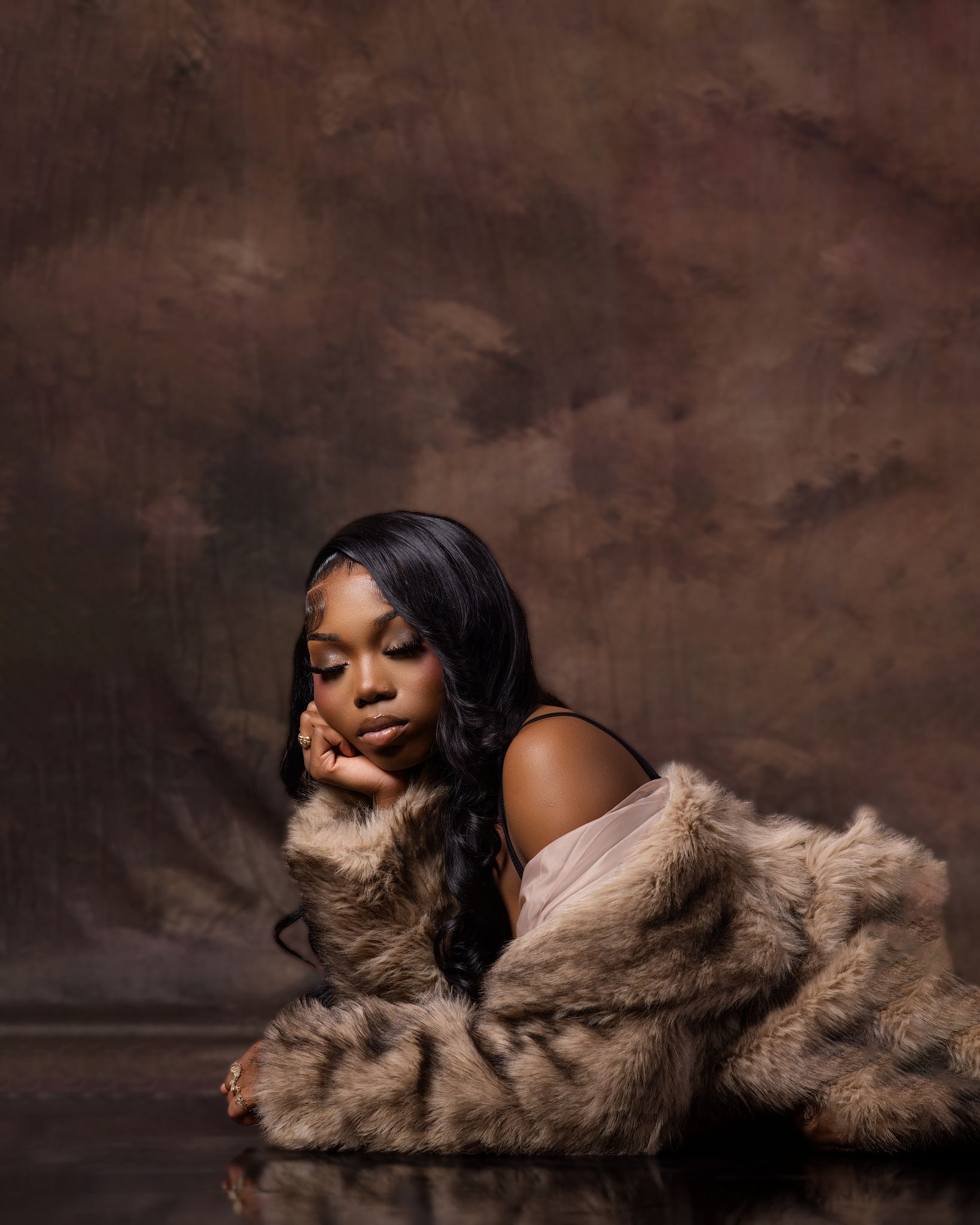 A young Black woman in a fur coat poses elegantly on a reflective floor against a brown textured studio backdrop.