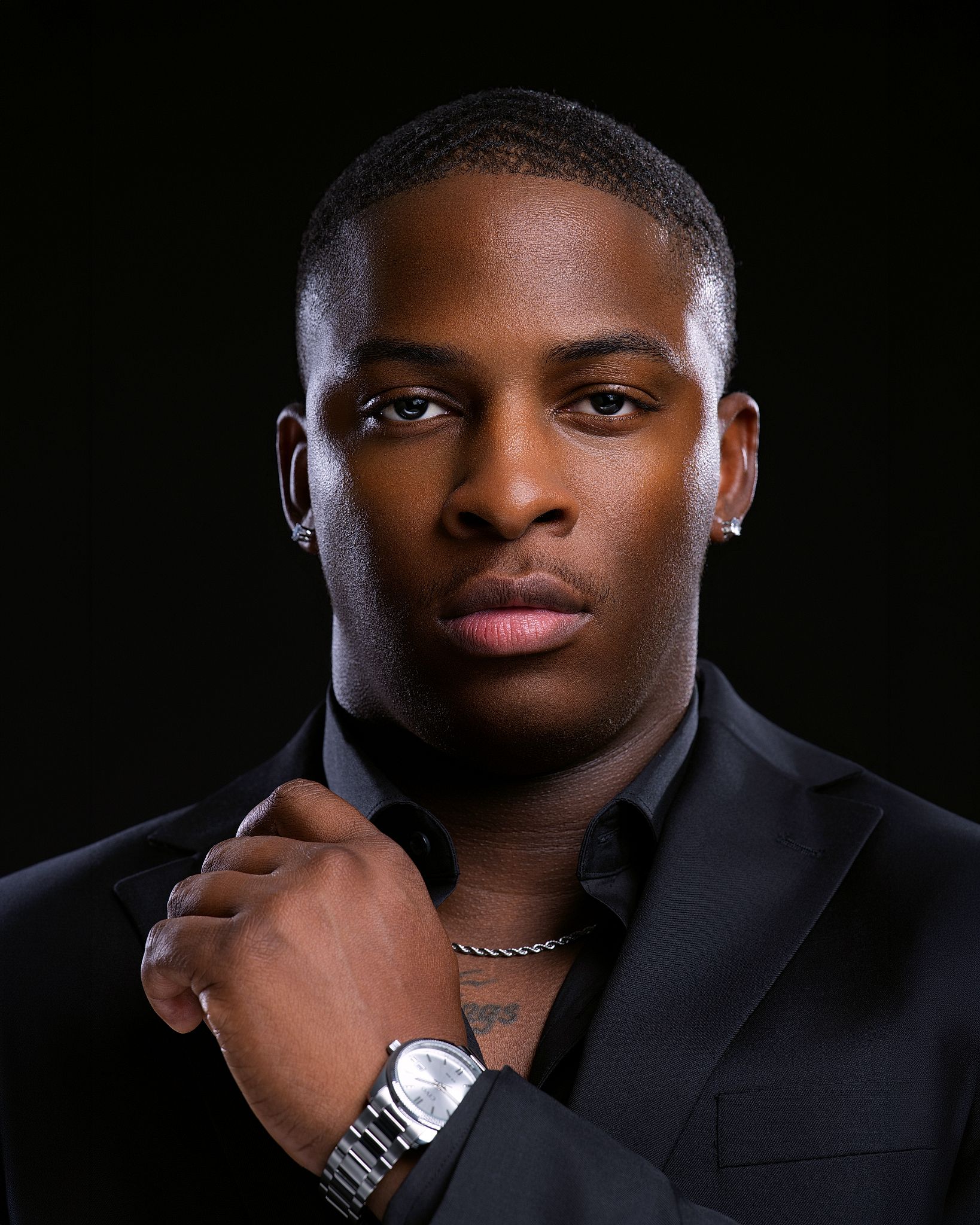 Young man in black suit adjusting collar, wearing silver watch and chain necklace, professional portrait photo.