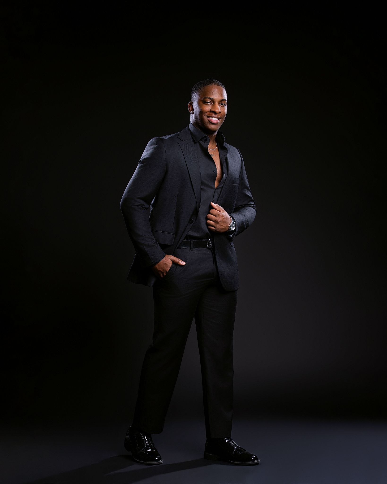 A confident man in a black suit poses against a dark background, smiling with one hand in his pocket.
