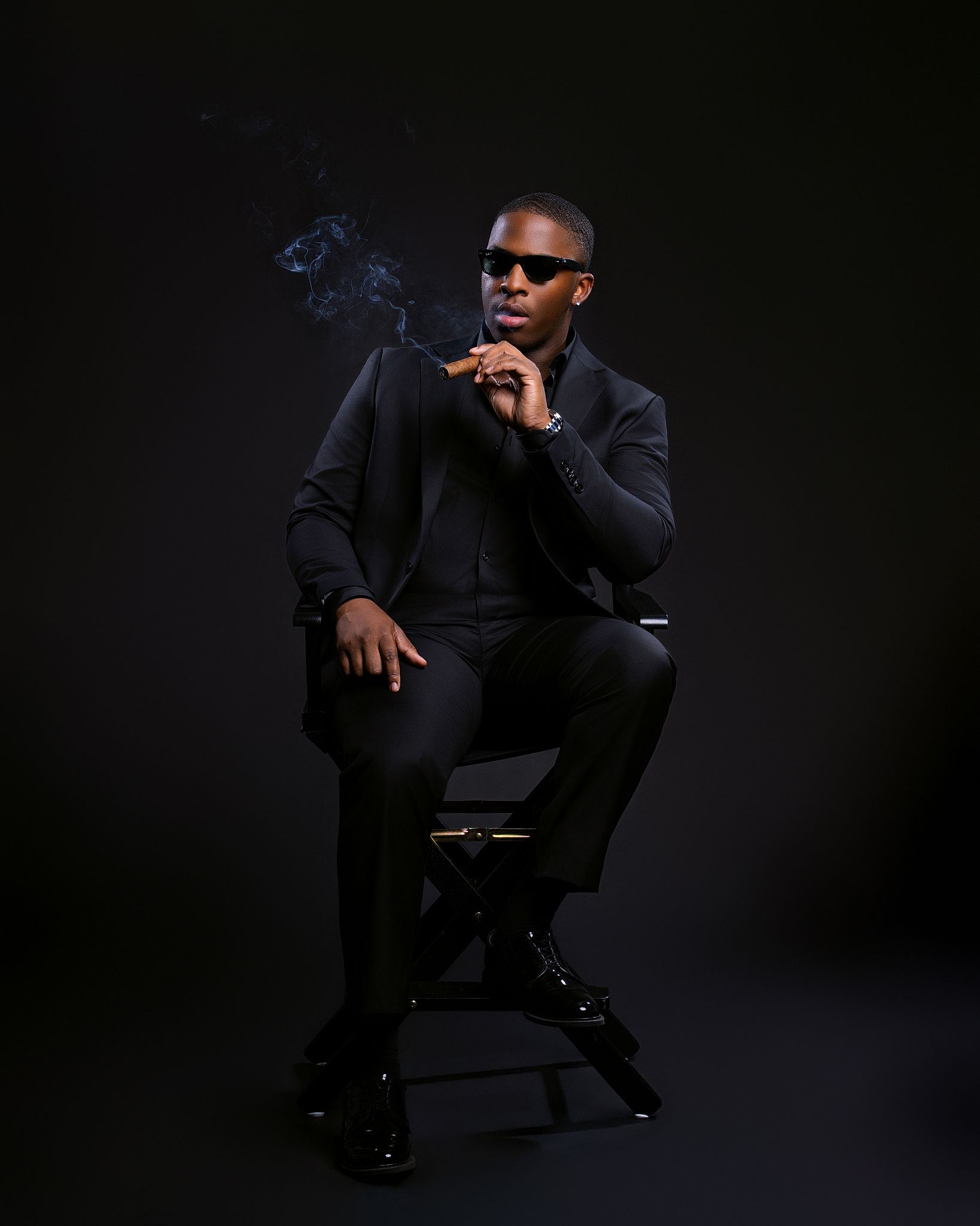 Man in black suit sitting on director's chair, smoking cigar, wearing sunglasses, dark studio background.