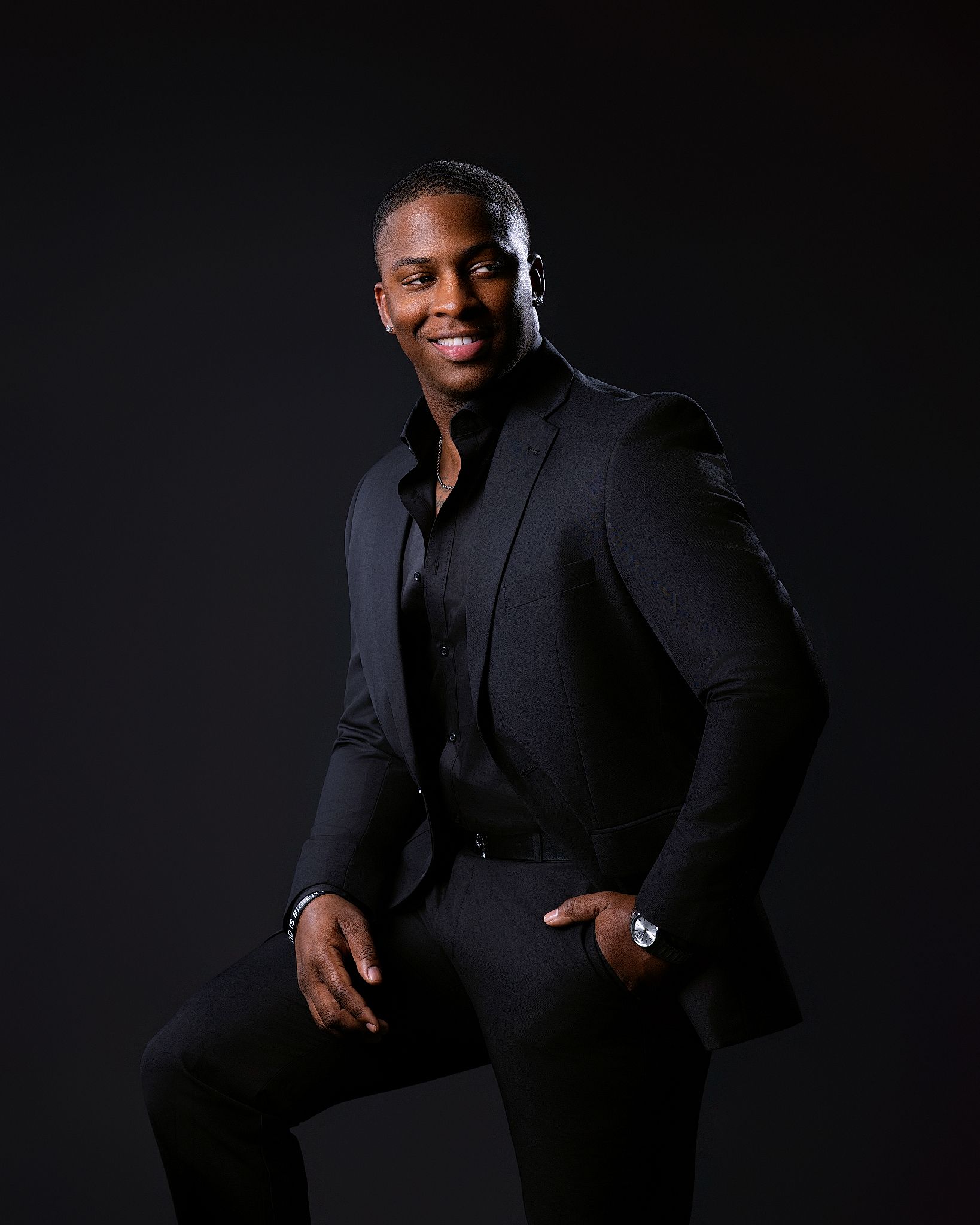 A confident man in an all-black suit poses against a dark background, smiling with one hand in his pocket.