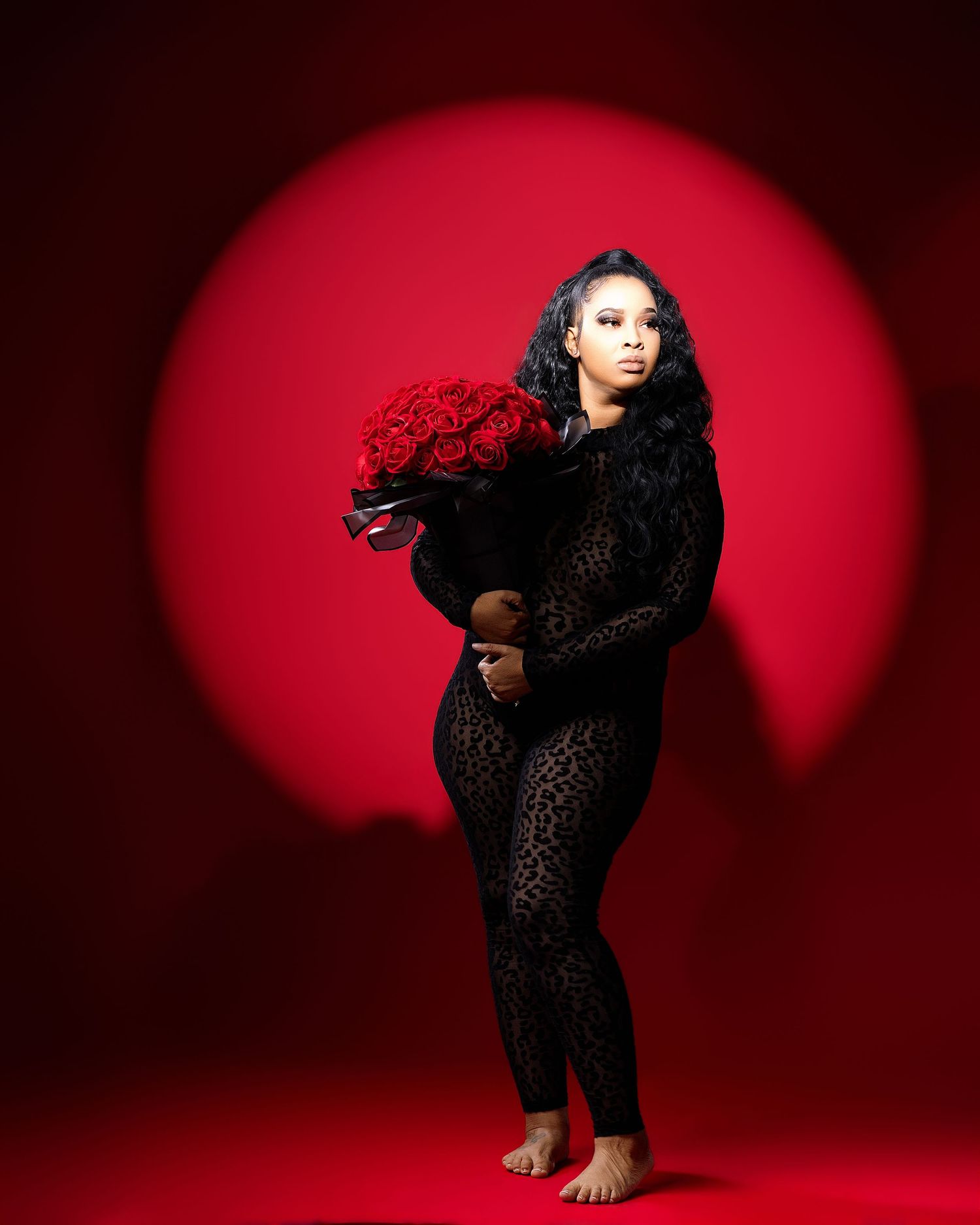 Woman in black lace jumpsuit holding red roses bouquet against vibrant red background with circular spotlight.
