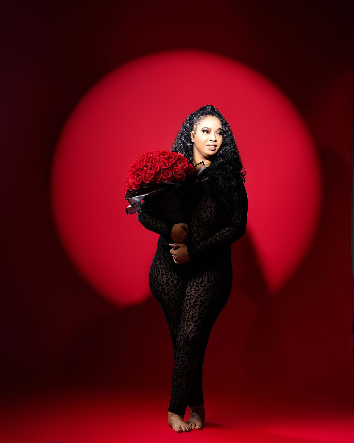 Woman in black lace jumpsuit holding red roses bouquet against vibrant red backdrop with circular spotlight.
