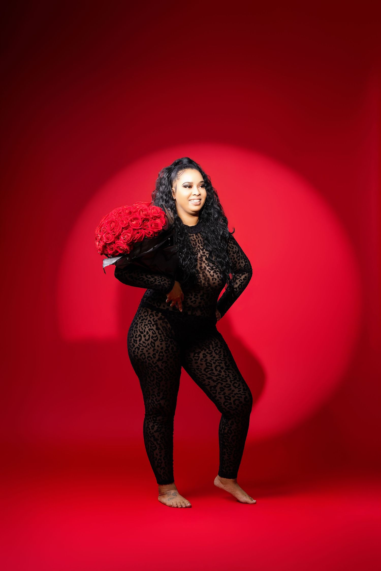 Woman in black lace jumpsuit holding red roses bouquet, posing against vibrant red studio background.