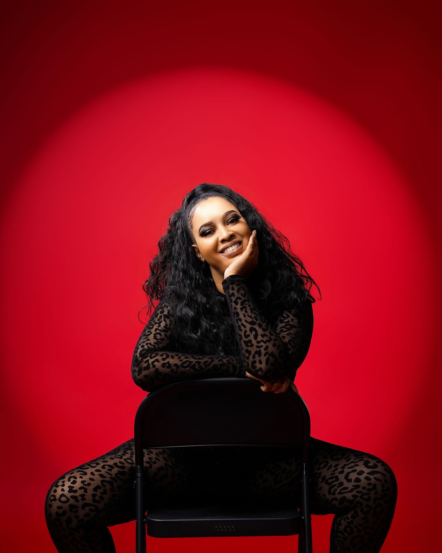 Woman in leopard print outfit poses on chair against vibrant red background, smiling confidently.