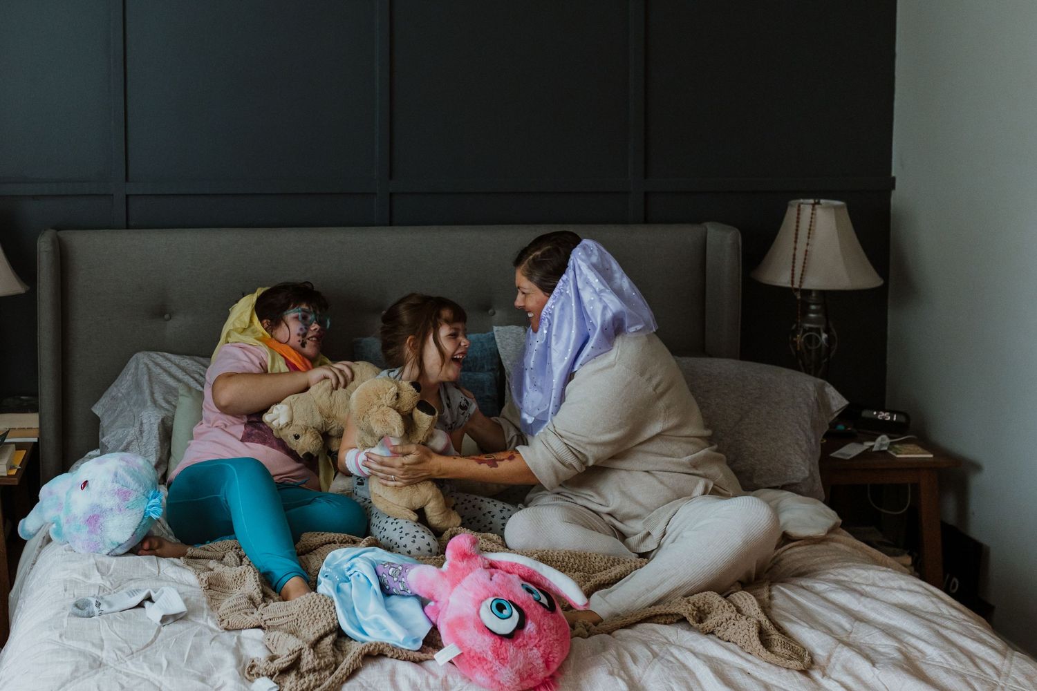 A mother and two children enjoy a cozy sleepover on a bed surrounded by stuffed animals and blankets.