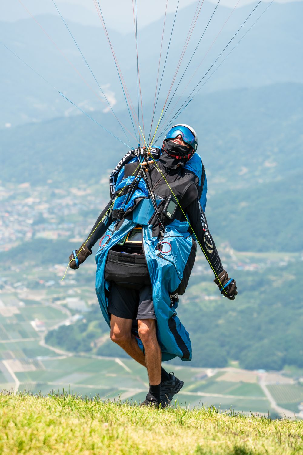 Paraglider in blue gear preparing for takeoff on a grassy mountain slope.