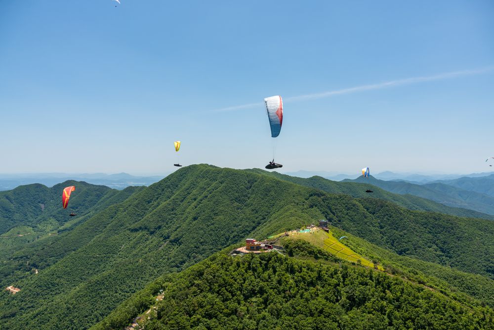 Multiple colorful paragliders soar over green mountain peaks against blue sky.