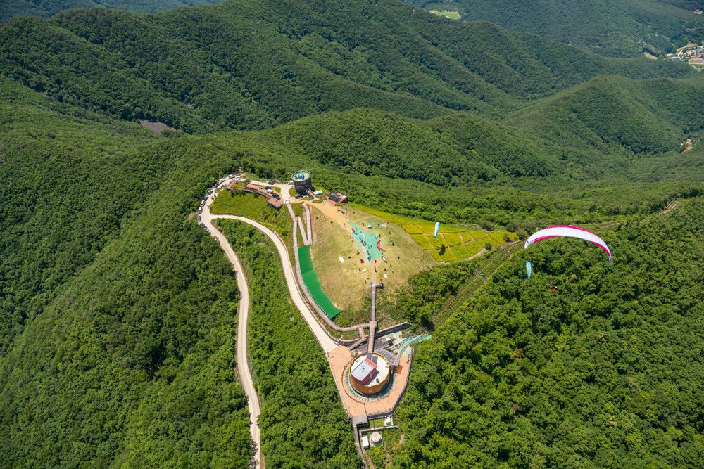 Aerial view of a paragliding launch site with winding paths surrounded by green mountains.