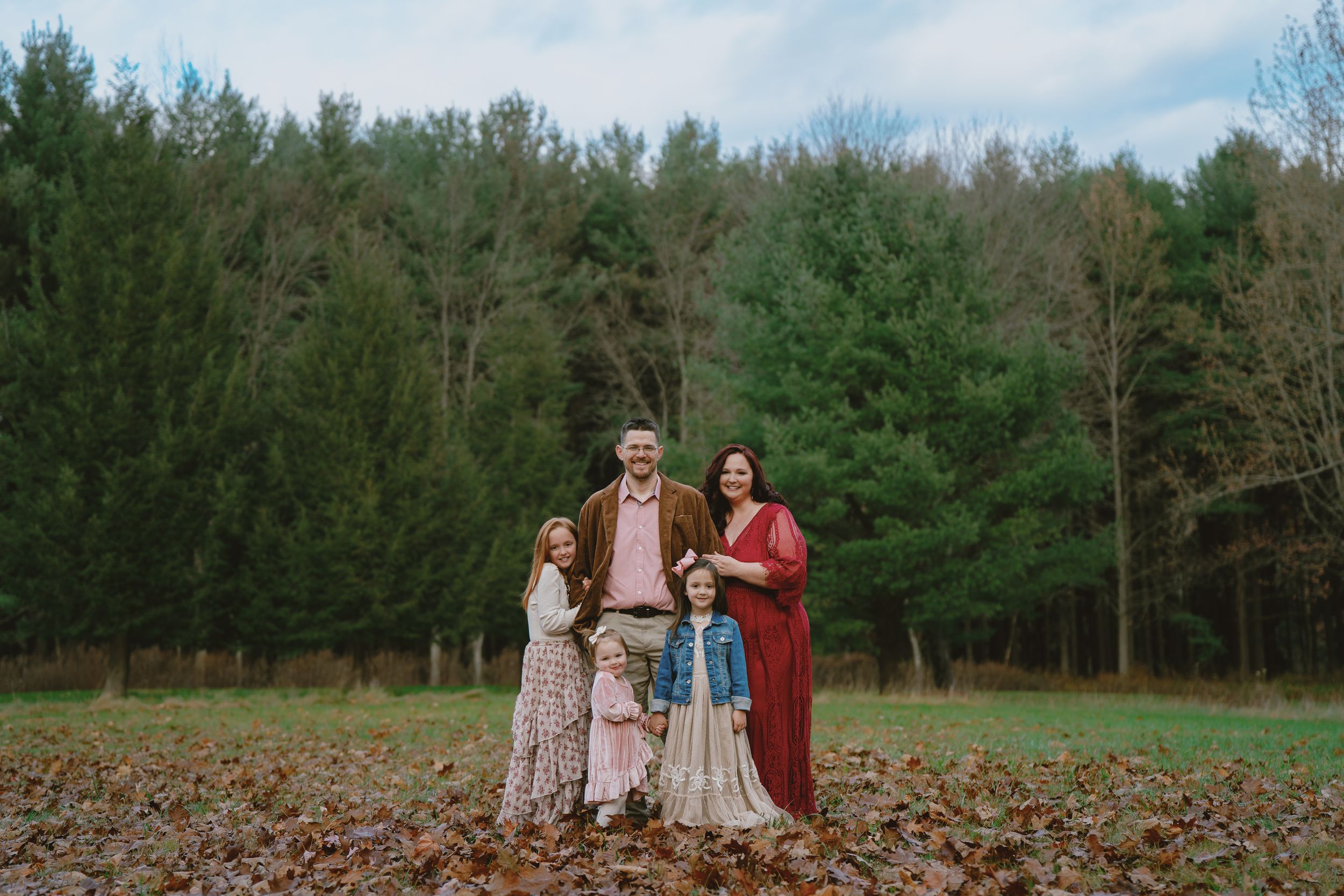 A family stands together in a field of fallen leaves with evergreen trees in the background on an autumn day in Cuyahoga Falls, Ohio for pic