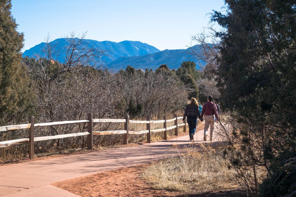 Paved trail with wooden fence leads toward distant blue mountains in Colorado.