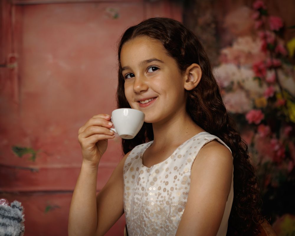 A young girl in a white lace dress sips from a small teacup against a pink floral backdrop.