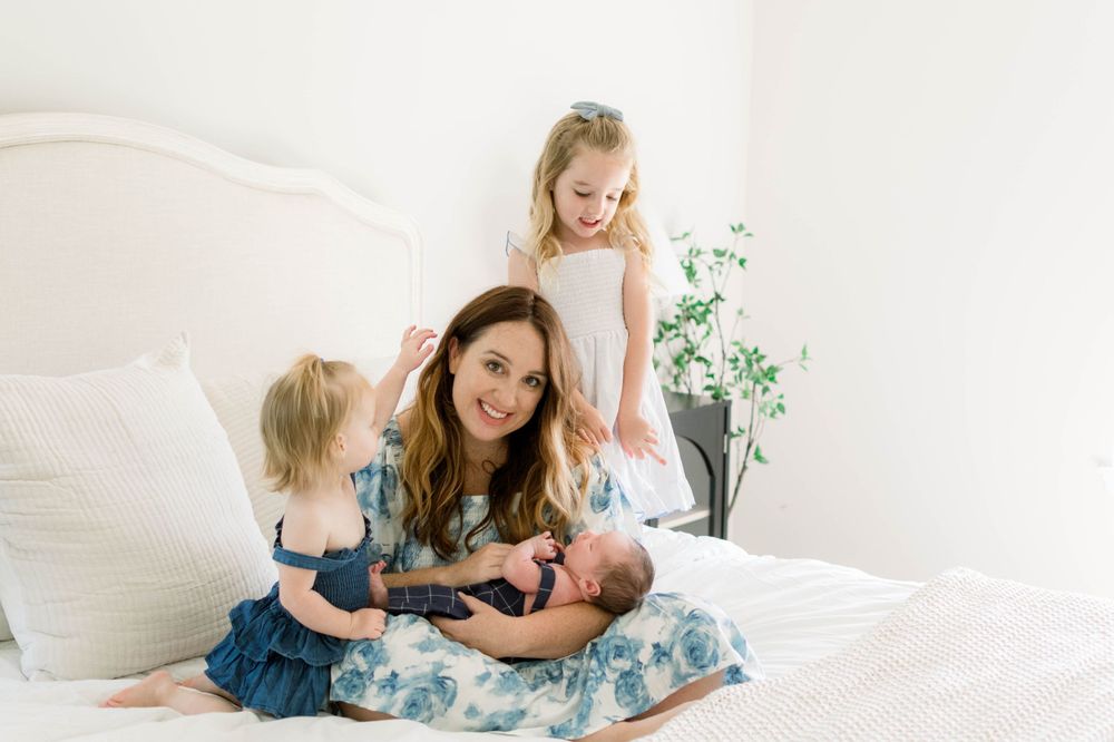A loving family moment captured on a white bed with a newborn baby and young children in soft natural light.