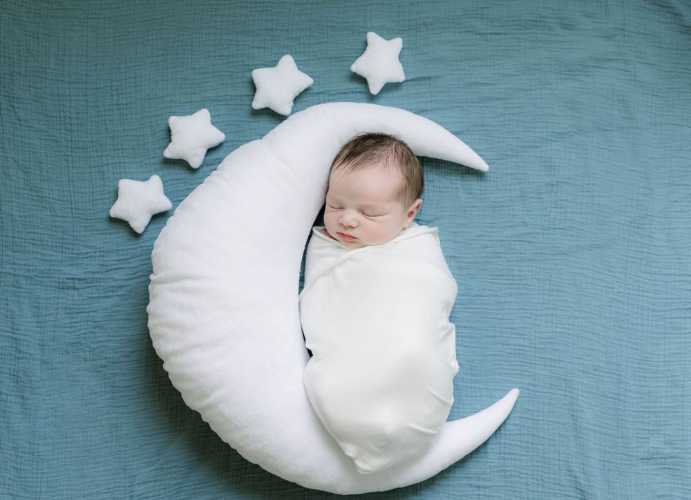 Swaddled newborn sleeping on a decorative white crescent moon cushion with stars.