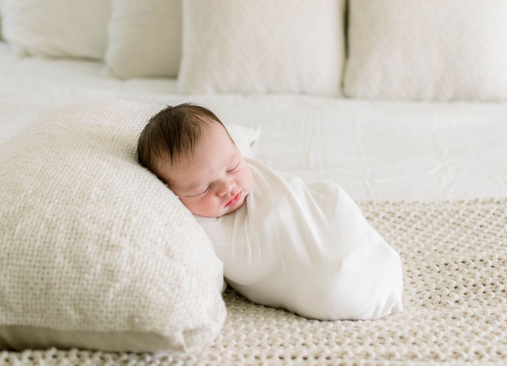 Newborn wrapped in white fabric resting peacefully against cream pillows.