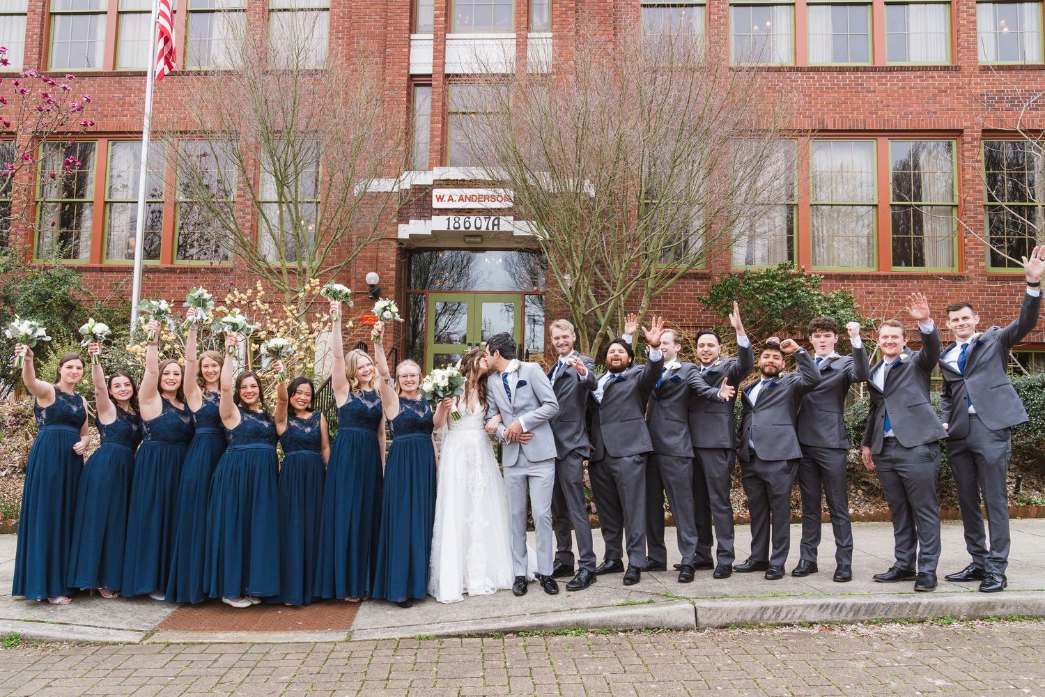 Wedding party lined up against brick building with bridesmaids in blue dresses and groomsmen in gray. McMenamin's in Bothell, Washington