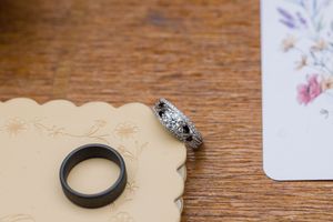 Two wedding rings, one black band and one diamond ring, rest on a decorative card beside floral stationery on wood.
