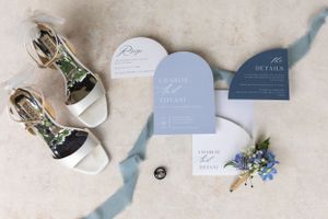 Wedding invitation suite with blue tones, sandals, and delicate floral accents on beige background.