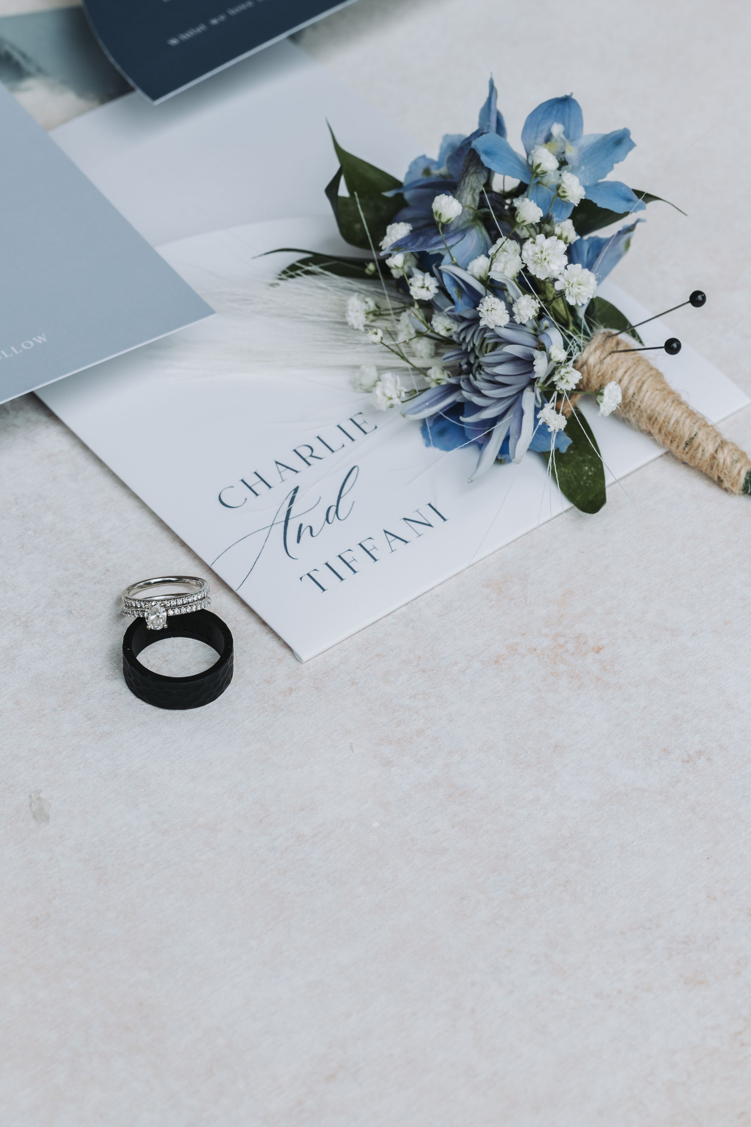 Wedding invitation with blue floral boutonniere and black ring on white surface.