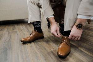 Groom ties brown dress shoes while wearing light khaki pants on hardwood floor.