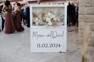 Wedding sign with floral decorations displays names and date 11.02.2024 against stone wall background.