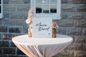 Elegant wedding signage on white tablecloth with dried floral arrangements against stone building.