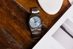 Silver wristwatch with light blue face displayed on wooden surface next to paper.