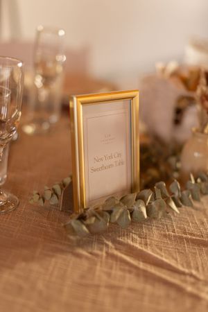 Gold-framed table number with eucalyptus leaves and champagne glasses on a textured tablecloth.