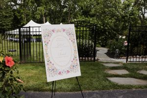 A floral welcome sign on an easel stands along a stone pathway near a black iron gate in an outdoor garden setting.
