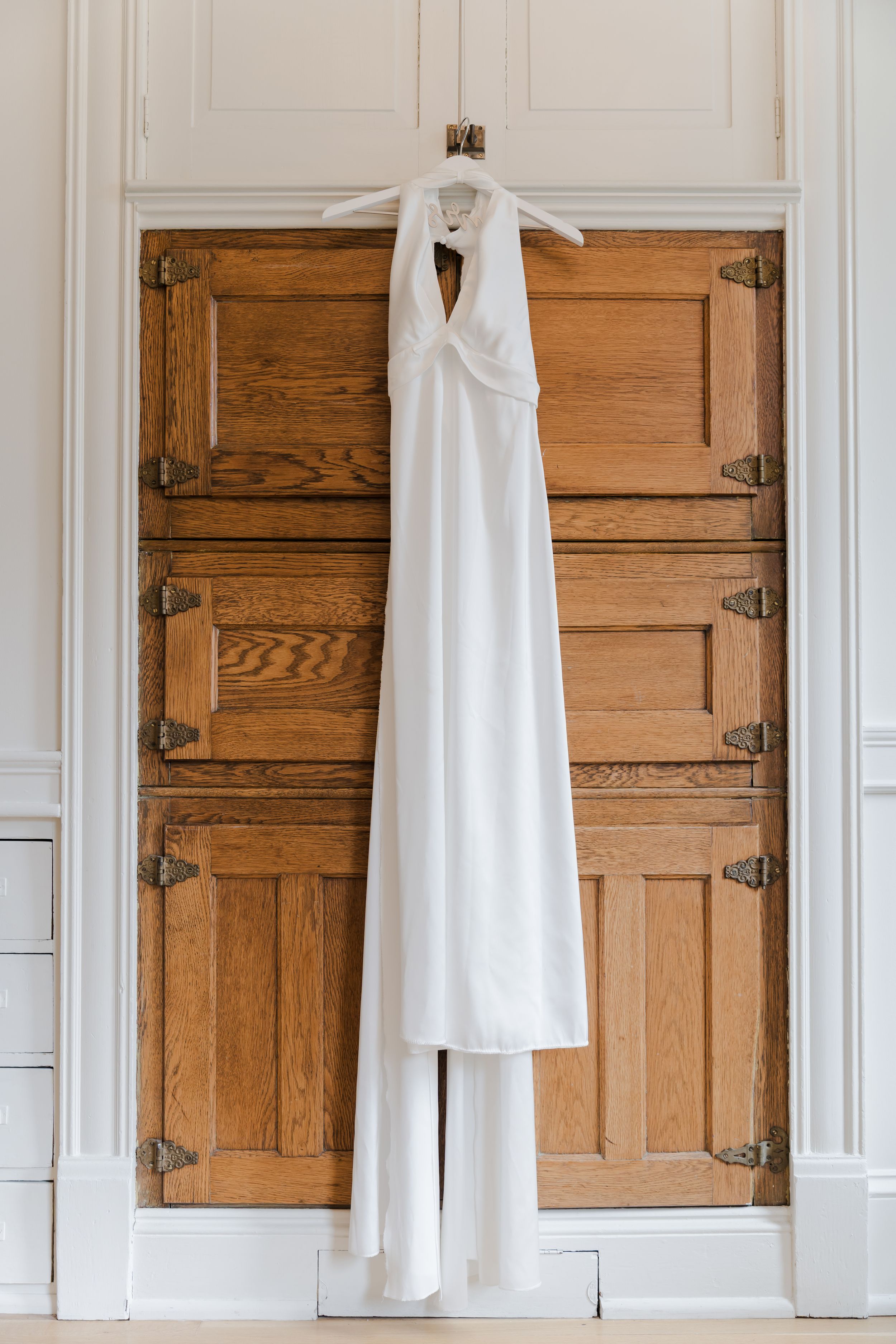Elegant white halter neck wedding dress hanging on wooden cabinet doors with brass hardware.