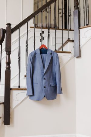 A light blue men's suit jacket hangs on a wooden staircase with dark iron balusters against a white wall.
