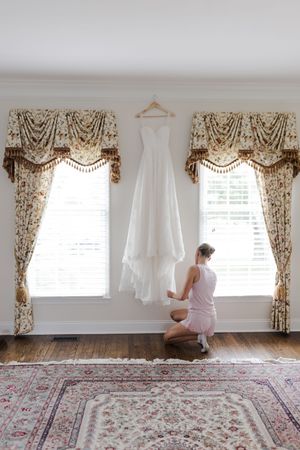 A white wedding dress hangs between elegant floral curtains in a bright room with hardwood floors and an ornate area rug.