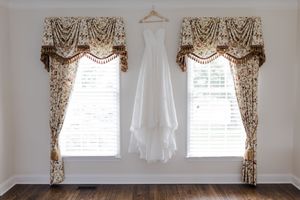 An elegant white wedding dress hangs between two ornate beige curtained windows in a sunlit room with hardwood floors.