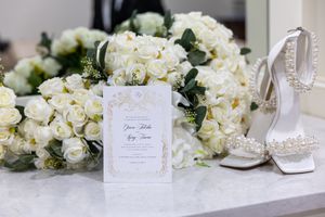 White wedding invitation card displayed with elegant bridal shoes and cream rose bouquets on marble surface.