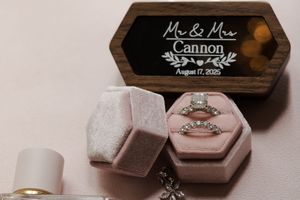 Wedding rings displayed on pink velvet hexagonal ring boxes next to rustic wooden name signs and decor.