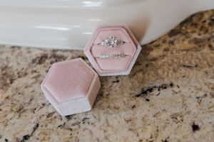 A sparkling engagement ring sits in a pink velvet hexagonal ring box on a marble countertop.