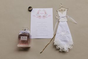 Elegant wedding details including perfume bottle, invitations, white dress, and accessories arranged on neutral beige background.