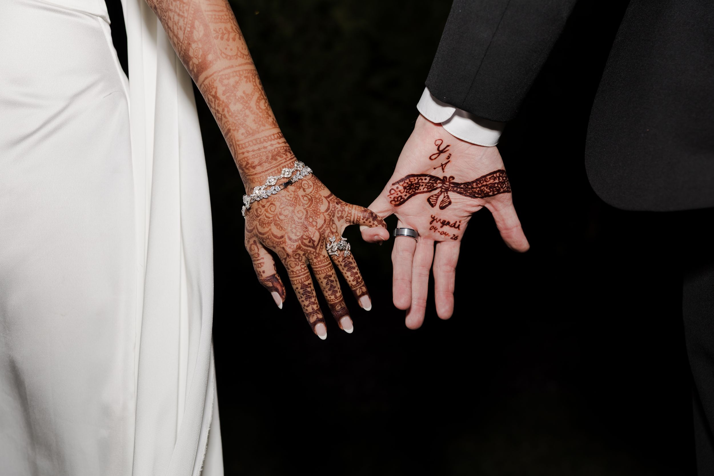 Close-up of two hands adorned with intricate henna mehndi designs, one in a white wedding dress, one in a black suit.