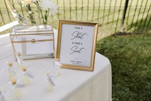 An elegant white table display features a gold-framed find your seat sign and clear acrylic card box at an outdoor wedding setup.